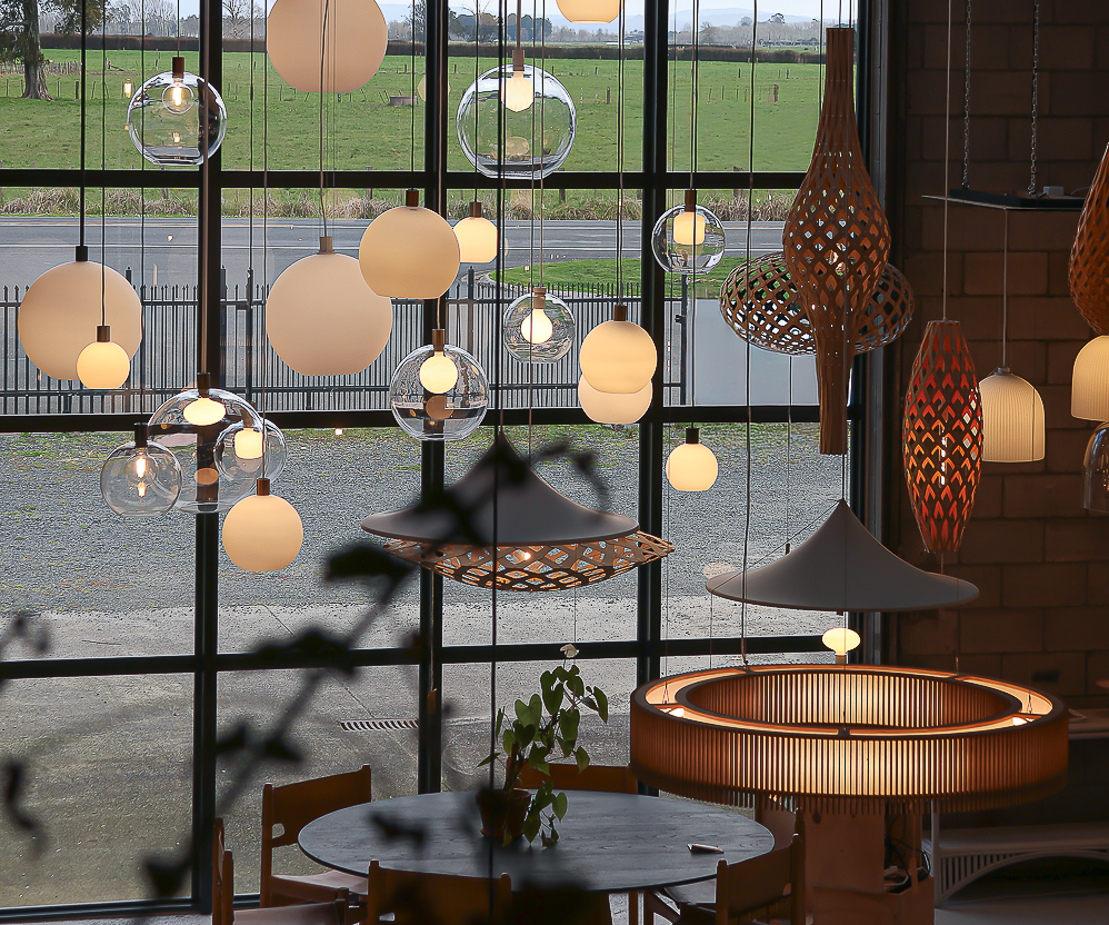 A collection of modern pendant lights in various shapes, sizes, and materials hanging in a large showroom with a window view of a green field.