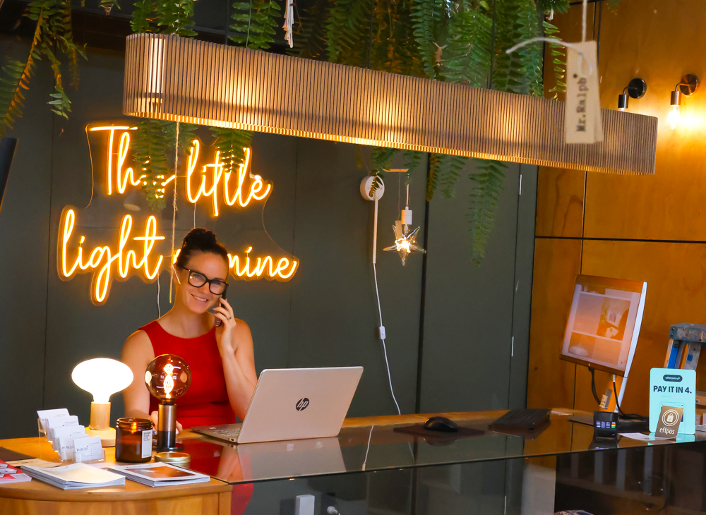 A woman in a red sleeveless top sits behind a wooden reception desk, working on a laptop in a modern cafe or restaurant setting. A glowing orange neon sign reads 'This little light mine' on the dark wall behind her.